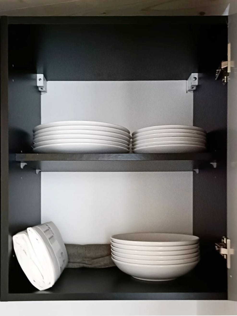 A kitchen cabinet is displayed with two shelves. The upper shelf contains several stacks of white plates, while the lower shelf holds white bowls and a gray kitchen towel neatly arranged. The interior is painted in a dark color, enhancing the cabinet's modern look.