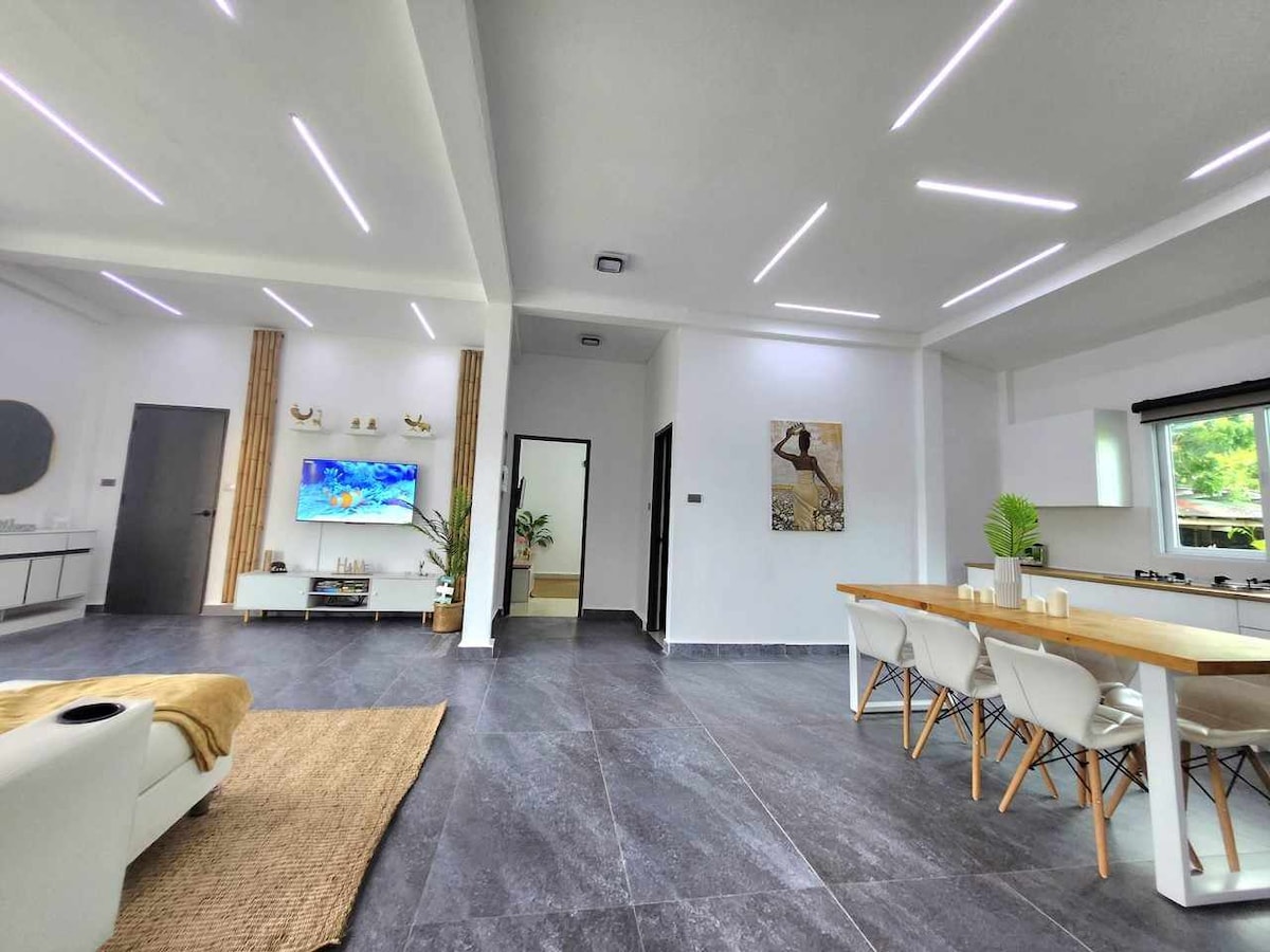 An open-concept living area features natural light streaming from large windows. A modern dining table with white chairs is visible, alongside a comfortable sofa. Soft lighting fixtures line the ceiling, and a large TV is mounted on the wall. Decorative touches enhance the space.