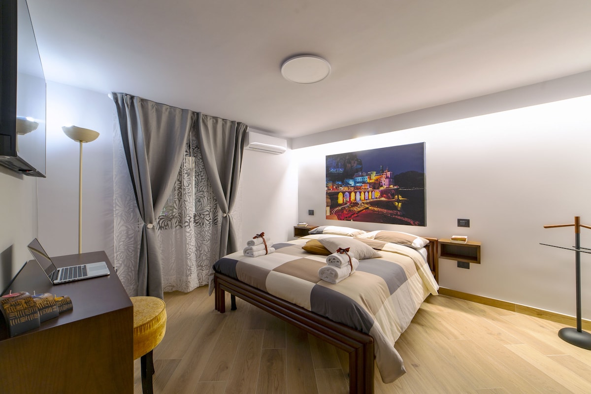 A well-appointed bedroom features a spacious bed with neutral-toned linens and decorative towels neatly arranged. A flat-screen TV is mounted on the wall opposite the bed. Soft lighting complements the modern decor, while a window dressed with sheer curtains allows natural light to filter in.