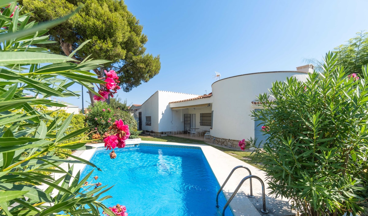 A private swimming pool is surrounded by lush greenery and colorful flowers, providing a serene outdoor space. The house features clean lines and a bright exterior, with large windows that invite natural light into the interiors. The clear blue sky complements the tranquil setting.