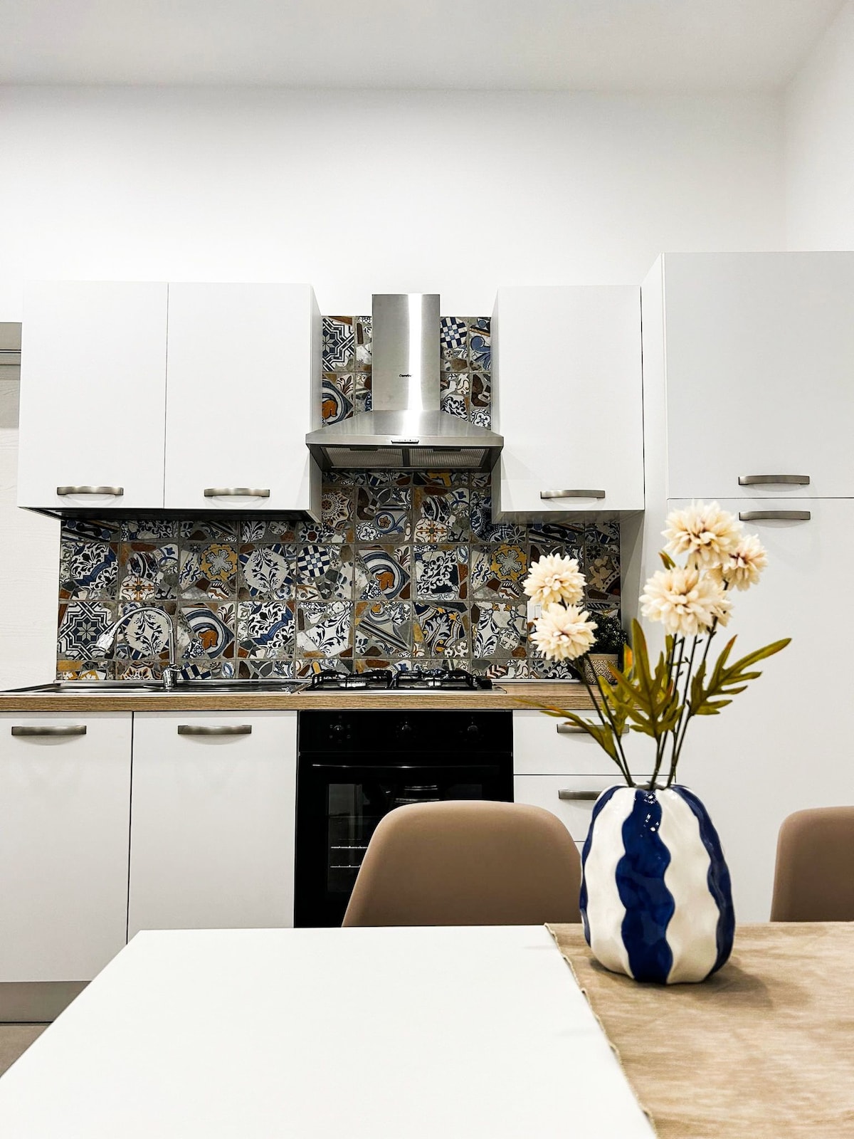 The kitchen features modern white cabinetry with a colorful tiled backsplash. A sleek range hood is mounted above the oven. A vase with flowers is displayed on a light-colored table, surrounded by neutral-toned chairs. Natural light illuminates the space.