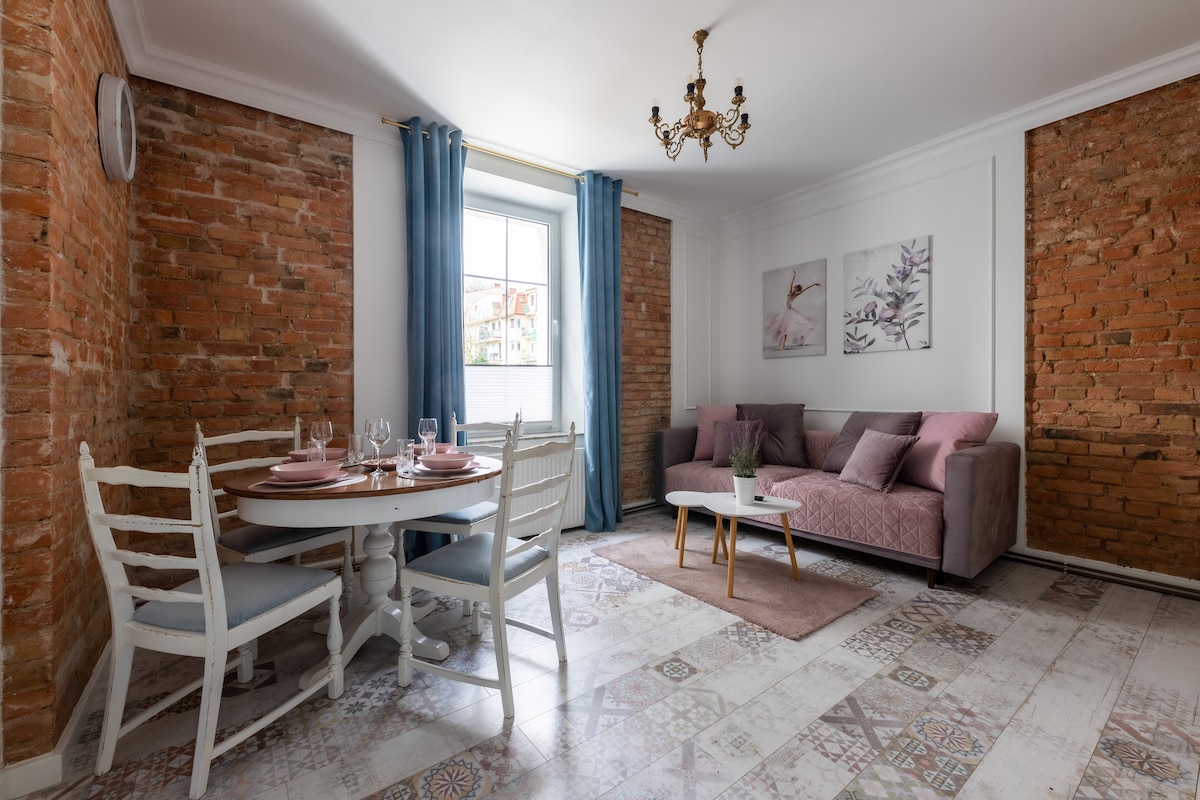 A bright living area features exposed brick walls and a mix of modern furnishings. A comfortable sofa and a round dining table set for four are present. Artwork hangs on the walls, and large windows allow natural light to fill the space.