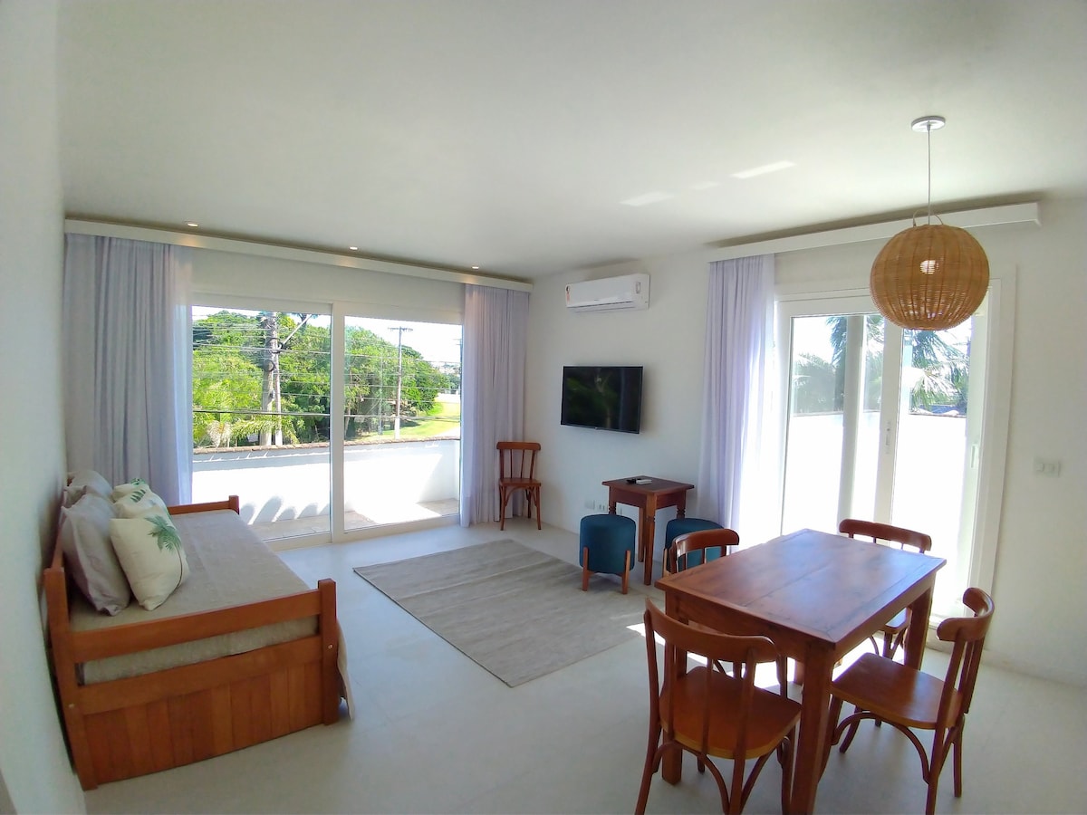 A bright living area features large windows allowing ample natural light and showcasing greenery outside. A wooden table with four chairs is positioned near the center, while a cozy seating area includes a daybed and a side table. A wall-mounted television adds convenience.