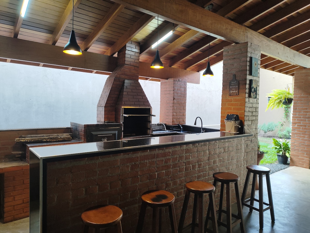 An outdoor kitchen area features a brick fireplace and grill, along with a long countertop surrounded by wooden stools. Pendant lights hang above, illuminating the space. Lush greenery is visible in the background, enhancing the connection to nature.