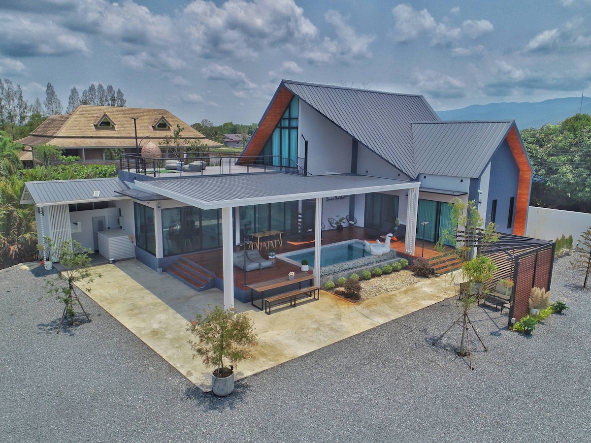 An aerial view of a modern lakeside retreat is presented, showcasing large outdoor spaces and a pool surrounded by landscaping. The structure features a combination of sleek lines and natural materials, with ample seating available on the spacious patio under a shaded area.