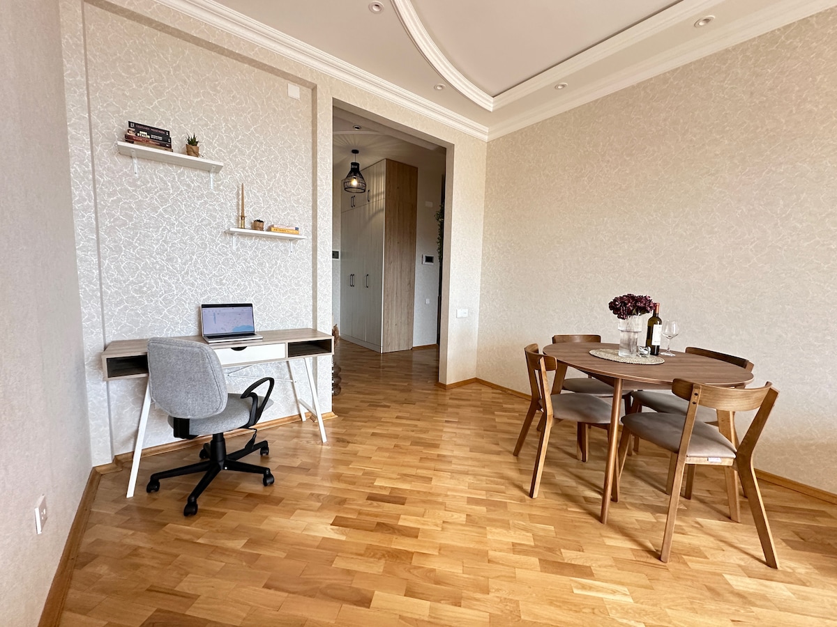 A well-lit dining area features a round wooden table accompanied by four chairs, creating a cozy setting. A workspace with a desk and chair is visible, complemented by a shelf displaying decorative items. The warm wood flooring adds to the inviting ambiance.