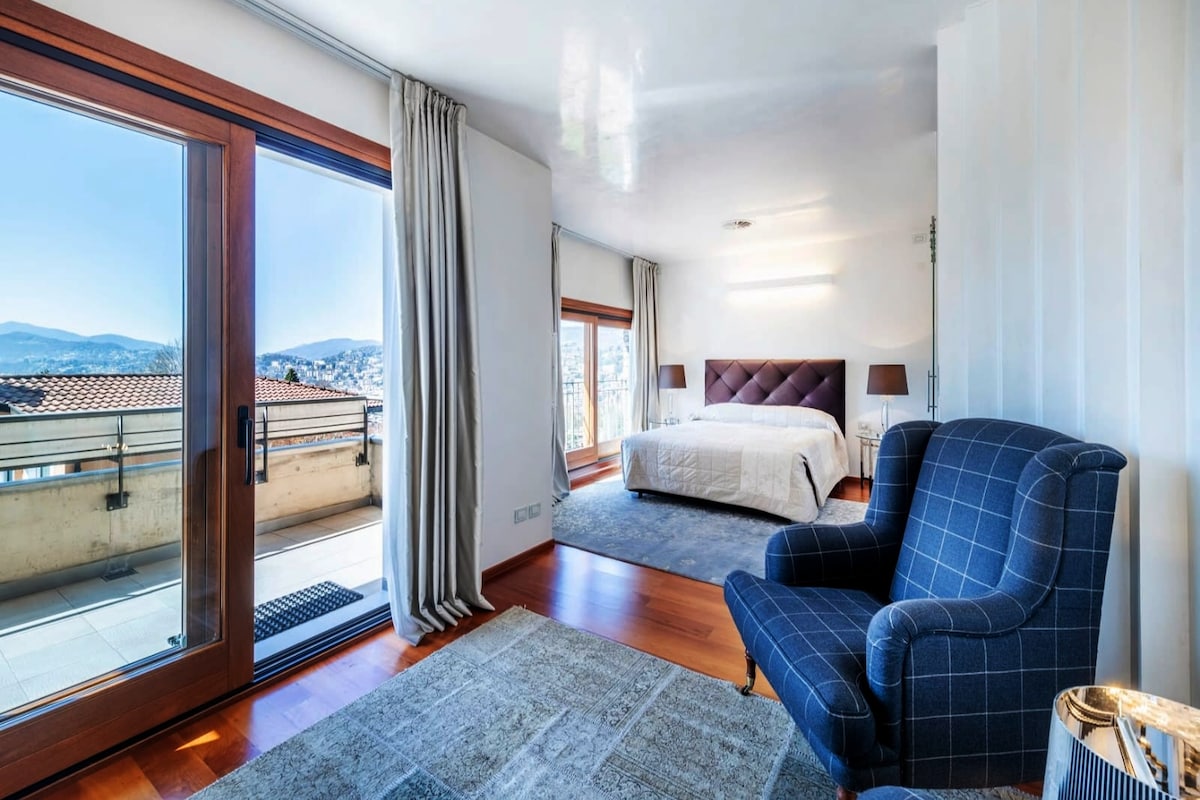 A bright bedroom features a spacious bed with an upholstered headboard and large windows that open to a balcony. A blue armchair is placed beside a small table on a soft, neutral rug, and natural light illuminates the room.