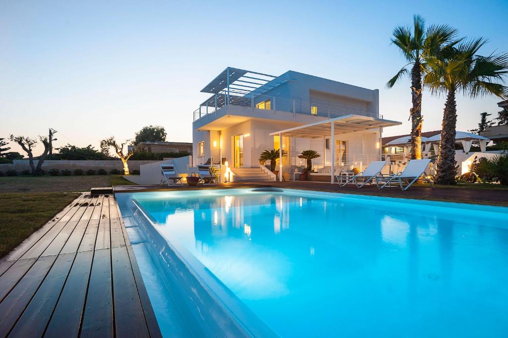 A modern villa is seen at dusk, with softly lit interiors reflecting on the tranquil swimming pool. Palm trees are positioned beside the pool area, and a wooden deck complements the outdoor space. The overall ambiance suggests a relaxing environment.