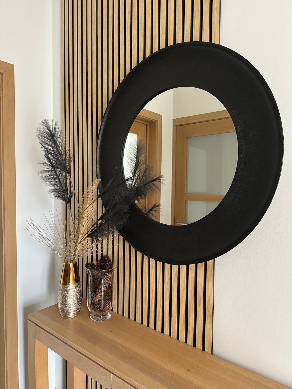 A round black mirror is mounted on a striped wooden wall, reflecting a warm interior. Below the mirror, a wooden console displays decorative items, including a vase with feathery accents and a glass container filled with natural elements. Soft natural light enriches the space.