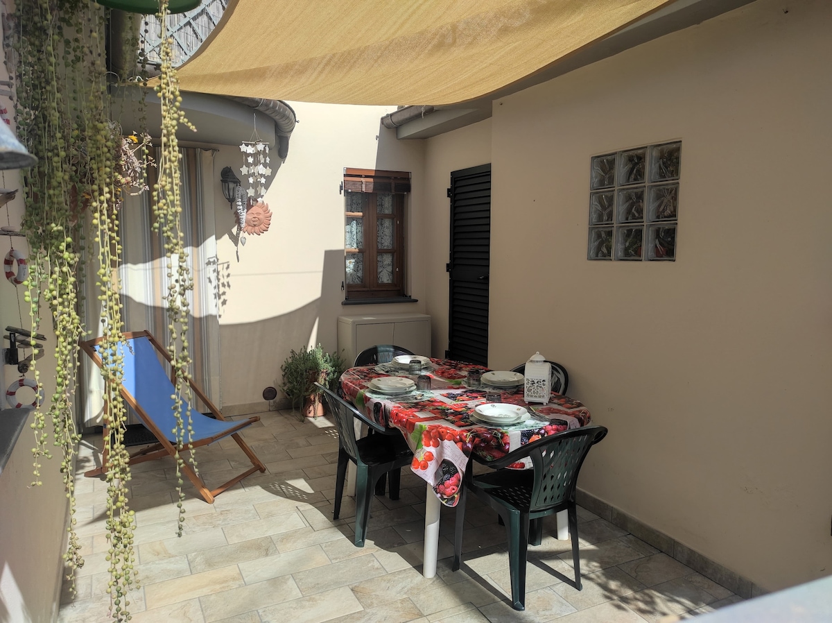 An outdoor dining area is highlighted with a table set for six, adorned with a floral tablecloth. Sun protection is provided by a canopy overhead. Surrounding plants add greenery to the stone-paved space, complemented by a comfortable lounge chair positioned nearby.