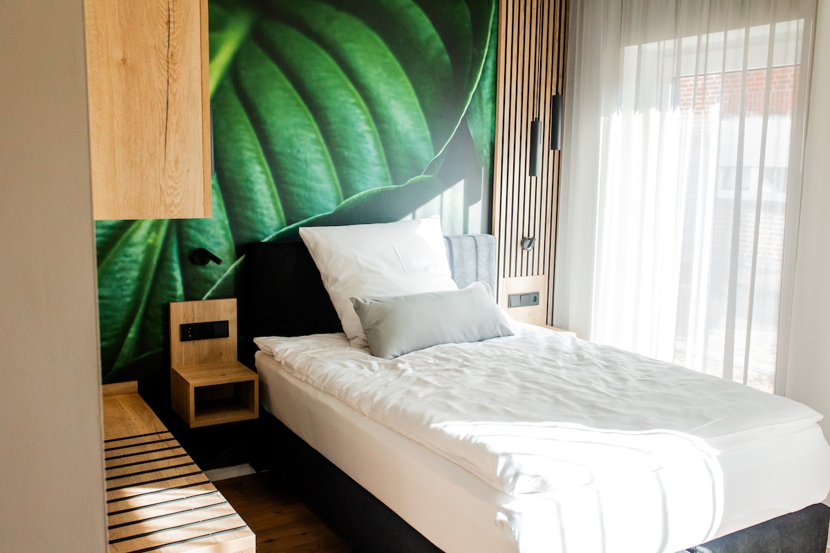 A sleeping area features a spacious bed adorned with crisp white linens and a decorative pillow. A tropical leaf mural adds a vibrant touch to the room. Natural light filters through sheer curtains, highlighting the modern wooden furnishings and textured wall elements.