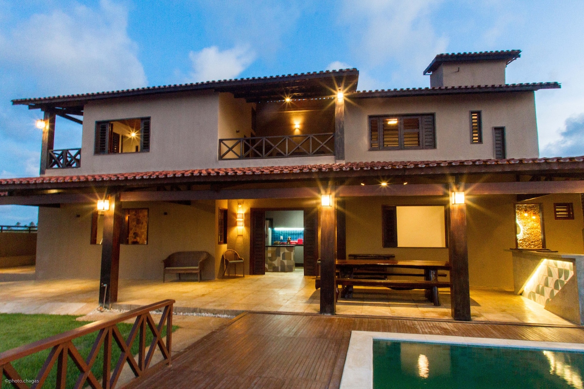 The two-story home features a welcoming facade with multiple outdoor seating areas, illuminated by soft lighting. A spacious balcony overlooks the garden, while large windows and doors provide a connection to the interior. A swimming pool and wooden deck enhance the outdoor space.