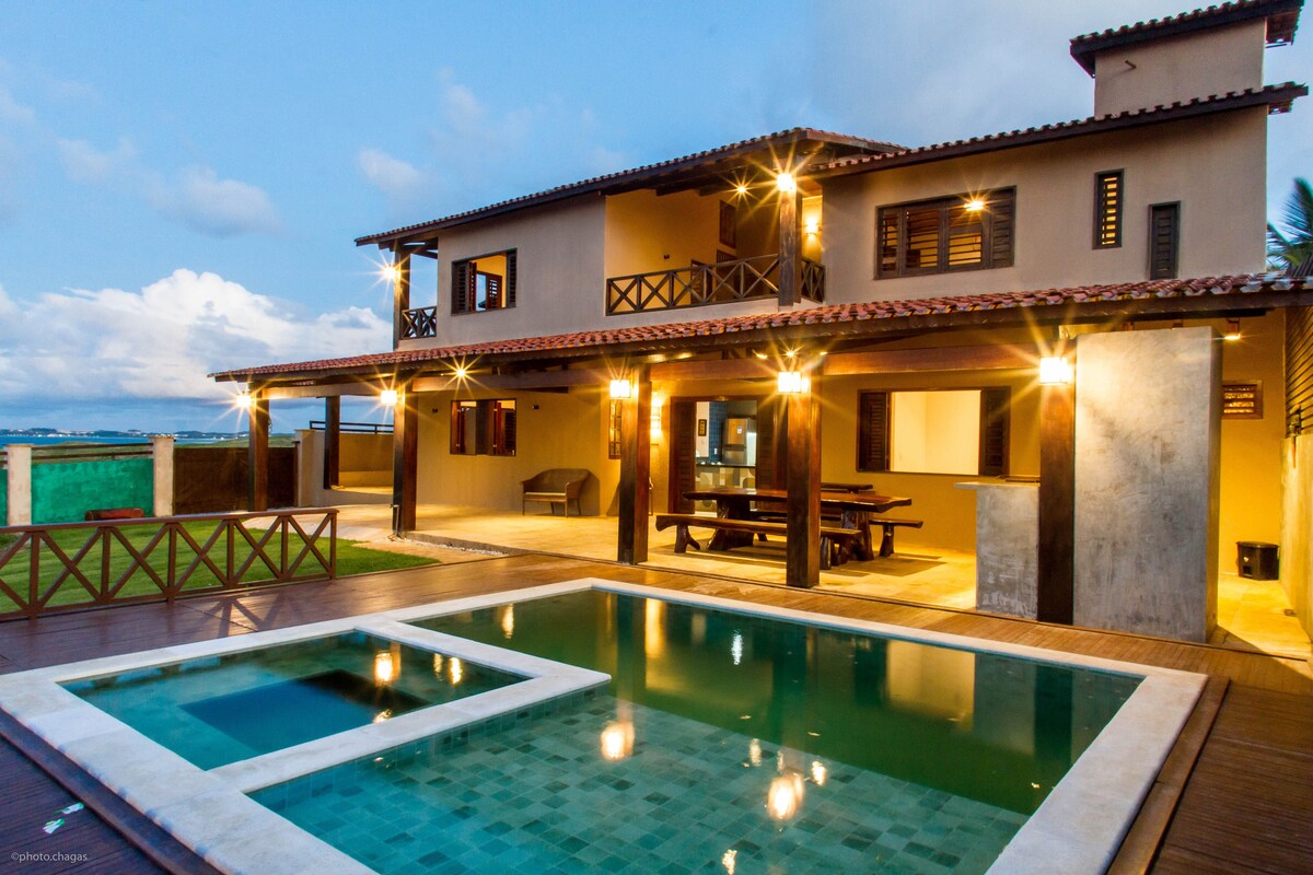 A spacious outdoor area features a swimming pool with a separate shallow section, set against a backdrop of a two-story house. Soft lighting illuminates the patio and surrounding gardens, highlighting the wood deck and natural stone walls.