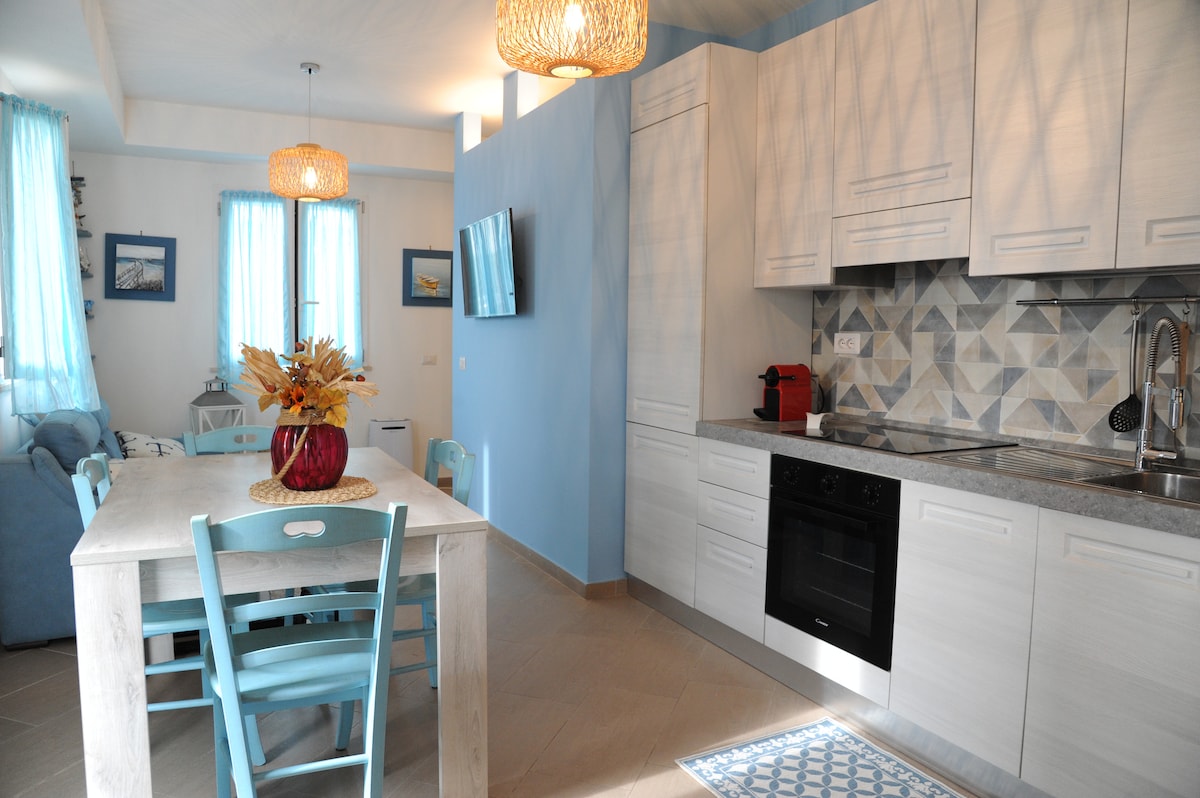 The living area features a modern kitchen equipped with a black oven, a sleek countertop, and a distinctive backsplash. A dining table with four blue chairs is accompanied by a decorative floral arrangement. Light streams in through the blue curtains, enhancing the airy ambiance.