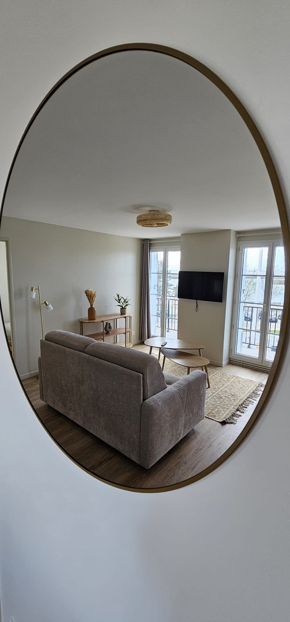 A living area is reflected in an oval mirror, featuring a neutral-colored sofa positioned against a wall. A wooden coffee table rests in front, accompanied by a decorative vase with greenery. Two large windows allow natural light to fill the space, enhancing the modern design.