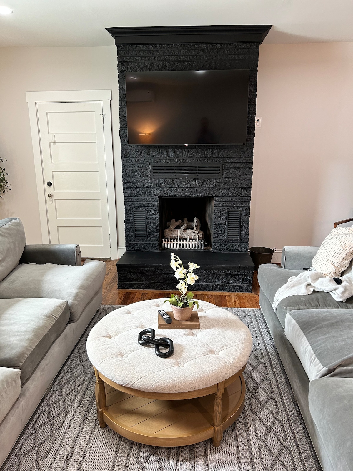 A cozy living area features a plush gray sofa facing a black stone fireplace with a mounted flat-screen television above. A round coffee table with a light-colored upholstered top is centered between the sofas, adorned with a small plant and decorative pieces.