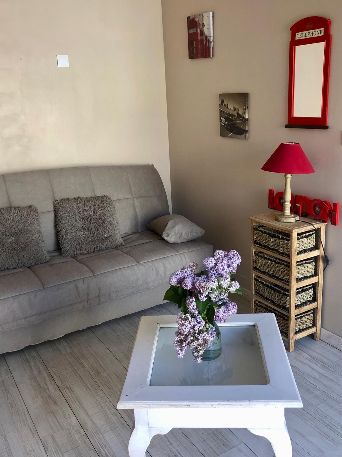 A cozy sitting area features a gray foldable sofa adorned with textured cushions. A small white coffee table with a vase of flowers sits at the center. Decorative items hang on the wall, while a red lamp adds a pop of color next to a woven storage unit.