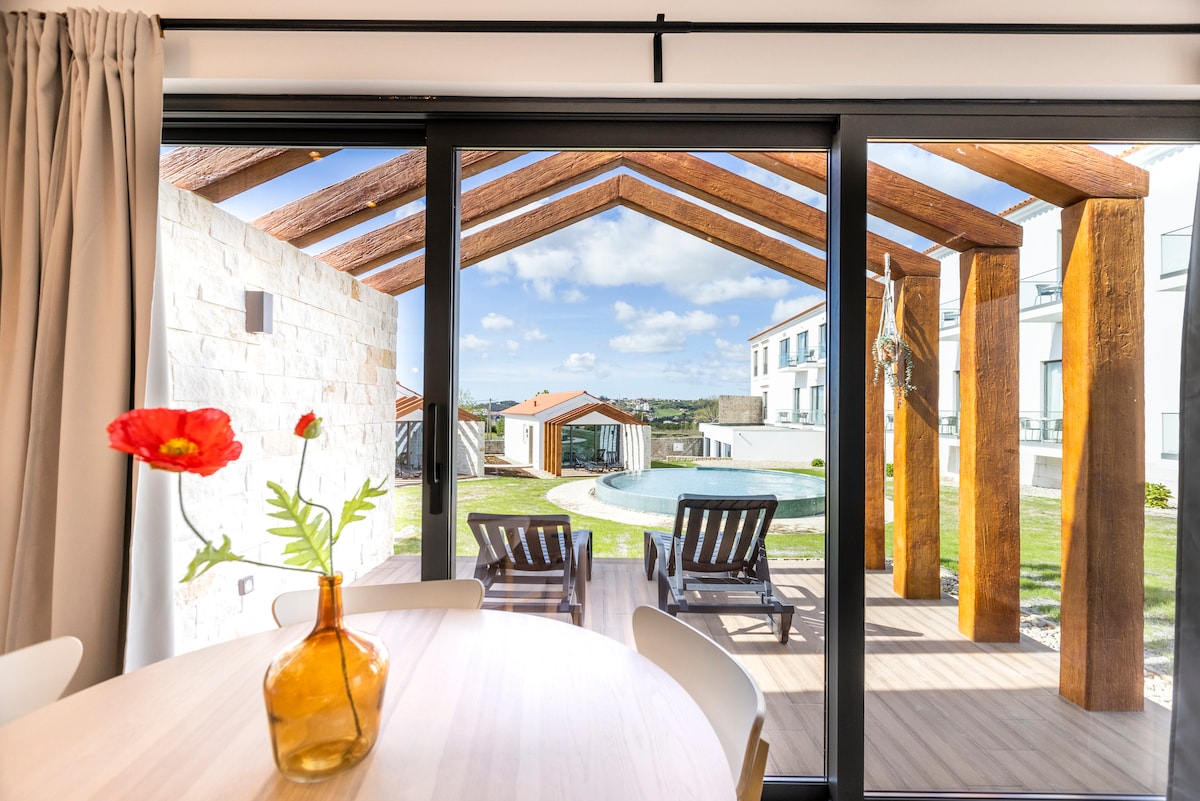 A dining area is complemented by a wooden table and a vase with a single flower. Large glass doors open to a patio area featuring wooden loungers and a pool, framed by a roof supported by wooden beams. Scenic views of the surrounding landscape are visible.