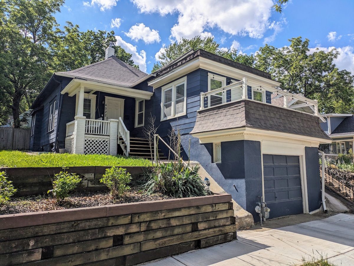 A charming 1920's bungalow is presented, featuring a welcoming porch with steps leading up to the entrance. The home showcases a blend of blue siding and natural landscaping, with a second-story balcony above the garage offering additional outdoor space.