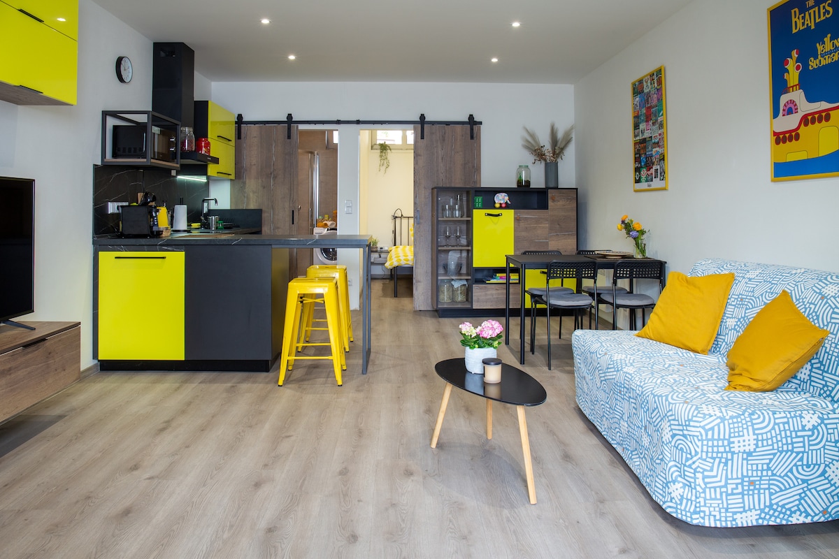 A modern living space is characterized by sleek cabinetry and bright yellow accents. A functional kitchen is equipped with appliances, while a cozy seating area features a patterned sofa and a coffee table. A dining area for four is visible, enhancing the open layout.
