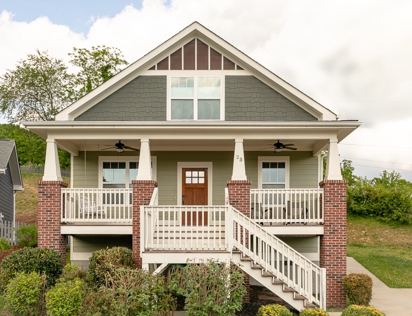 A well-crafted two-story home features a charming front porch with white railings and steps leading to the entrance. The exterior showcases a combination of grey siding and brick accents, with green landscaping framing the base. Clear windows invite natural light into the inviting space.