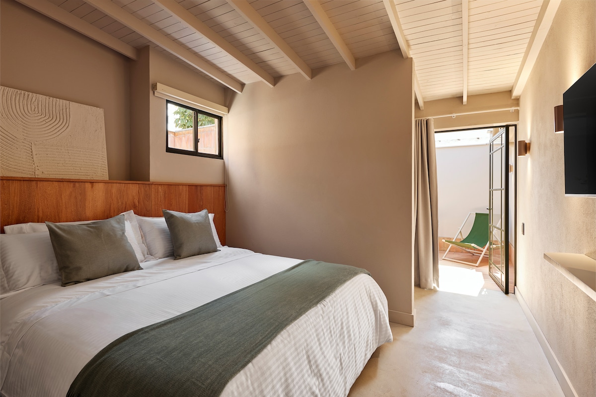 A spacious bedroom is showcased, featuring a double bed adorned with decorative pillows and a textured throw blanket. Natural light filters through a window, illuminating the neutral-toned walls. An open door reveals a private terrace, enhancing the room's connection to outdoor space.