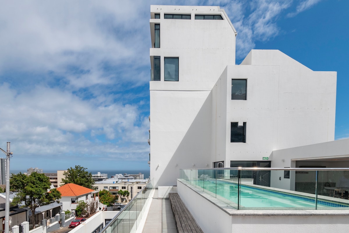The modern building structure displays a sleek, minimalist design with large windows and multiple levels. A spacious pool deck is visible in the foreground, featuring a shimmering pool and views of the surrounding neighborhood and the ocean in the distance.