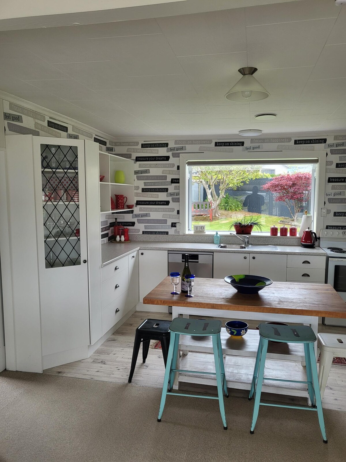 A well-equipped kitchen features white cabinetry with glass panel accents. A dining table made of wood sits in the center, surrounded by stools in light blue. Natural light enters through a large window, offering a view of the garden and nearby vibrant foliage.