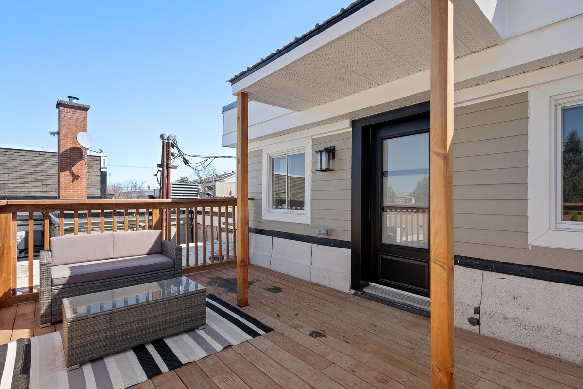An outdoor patio area features lounge seating with a coffee table, all situated on wooden decking. A striped outdoor rug adds a decorative touch. The area is surrounded by a railing, providing a view of the surroundings and access to the entrance of the unit.