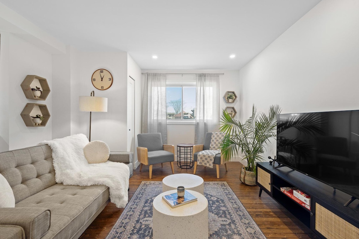 A comfortable living area features a grey sectional sofa adorned with a white throw blanket. Two accent chairs provide additional seating beside a modern coffee table. A large flat-screen TV rests on a black media console, while natural light pours in through a window dressed with sheer curtains.