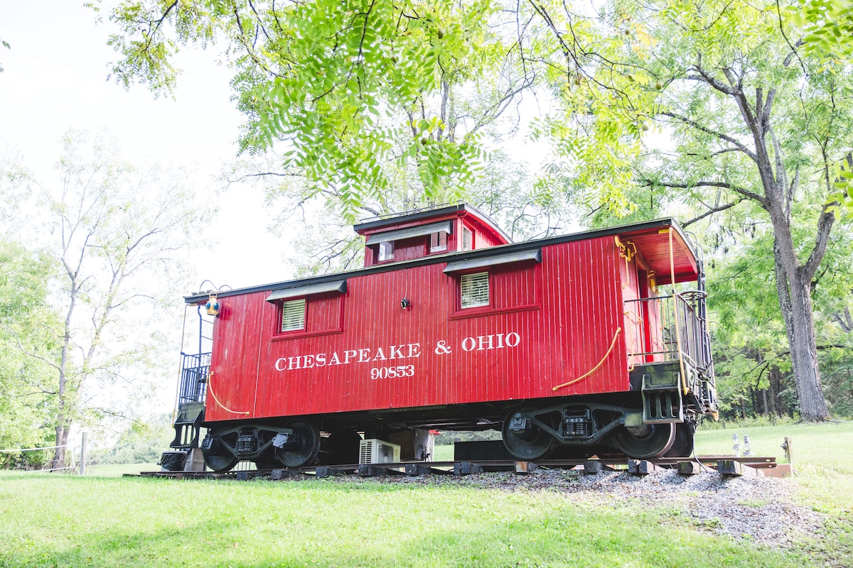 A vibrant red caboose is set among lush greenery, with a traditional design that emphasizes its historical significance. The structure features black wheels and a cupola on top, blending into the natural surroundings while offering a glimpse of its charming character.