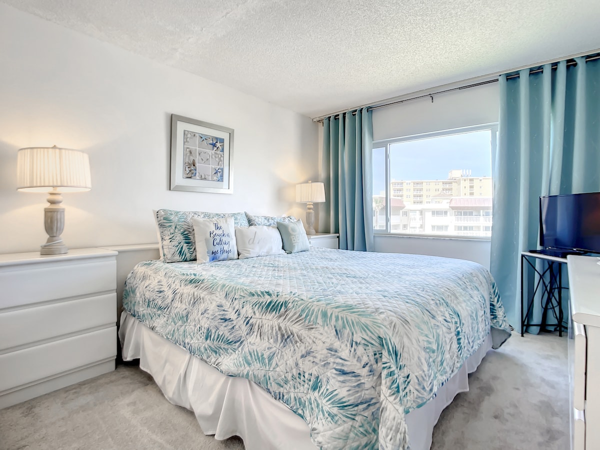 A spacious bedroom features a large bed dressed in a tropical-patterned quilt. Soft blue curtains frame the window, providing natural light. A dresser with lamps and a small TV on a white desk add functionality to the inviting space.
