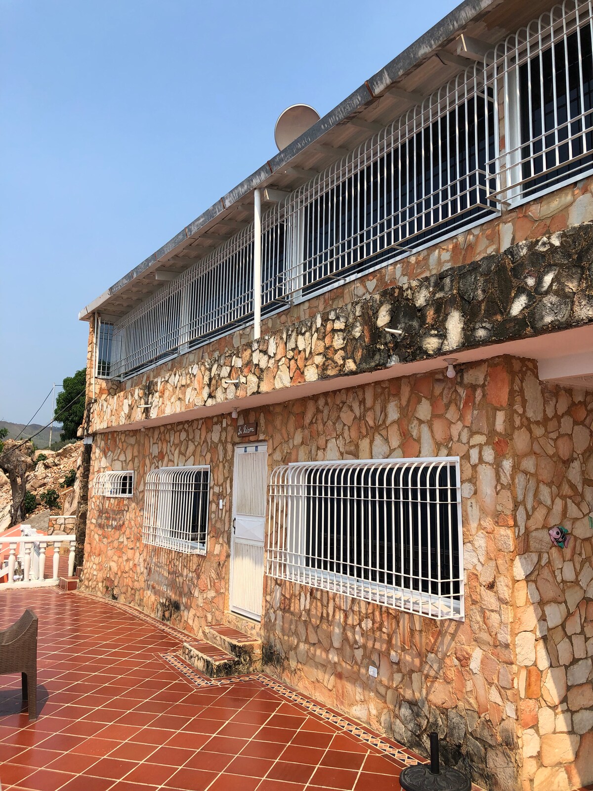 The exterior of a stone-faced building is highlighted, showcasing a spacious patio with red-tiled flooring. Windows are secured with metal bars, and a door is centrally located. The structure's upper level features additional metal-barred windows, reflecting local architectural style.