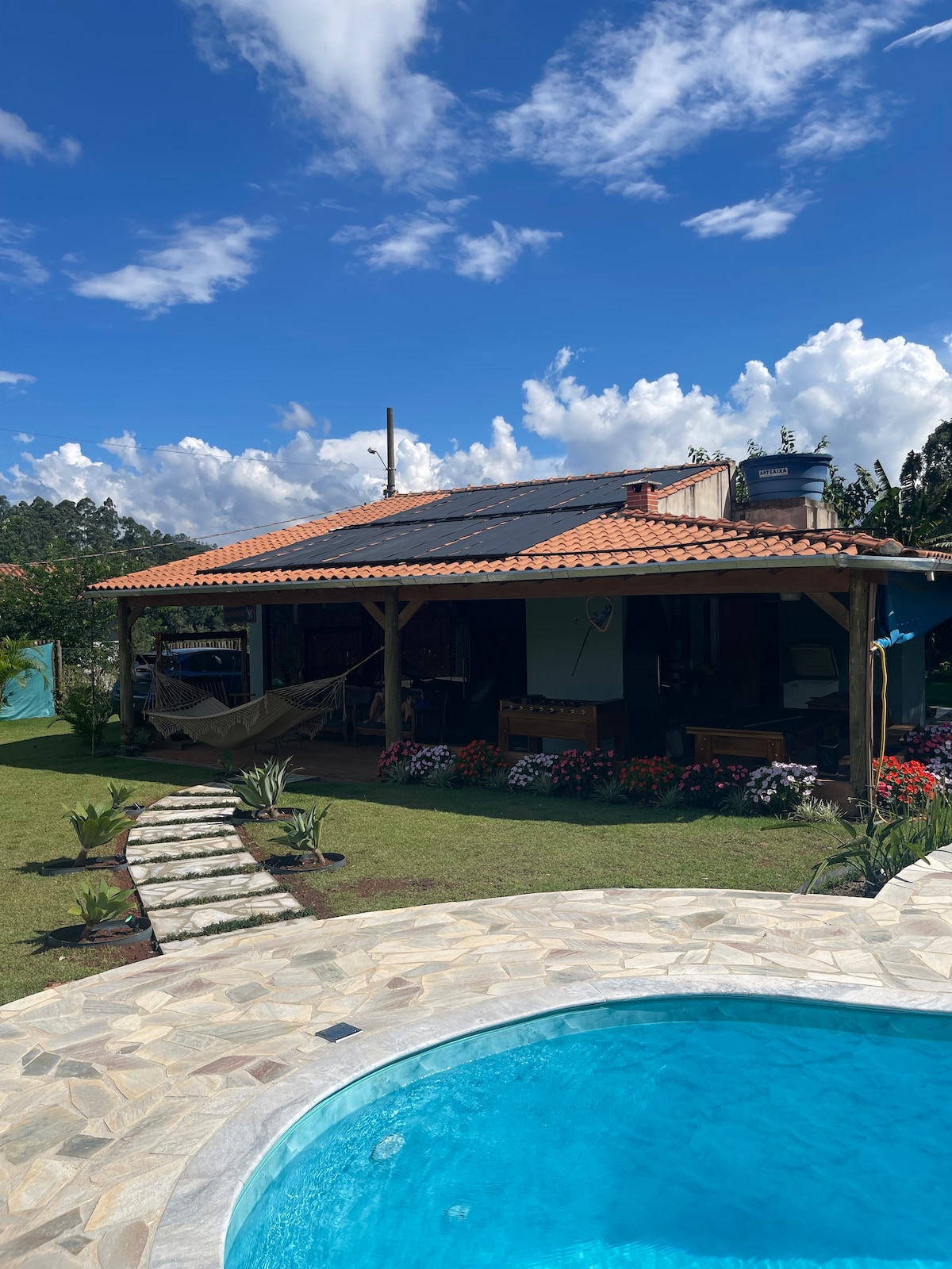 A charming house with a terracotta roof sits amidst vibrant gardens, featuring a pathway made of stone leading to a pool. The inviting outdoor area is complemented by greenery and a shaded porch, providing a cozy retreat surrounded by nature.