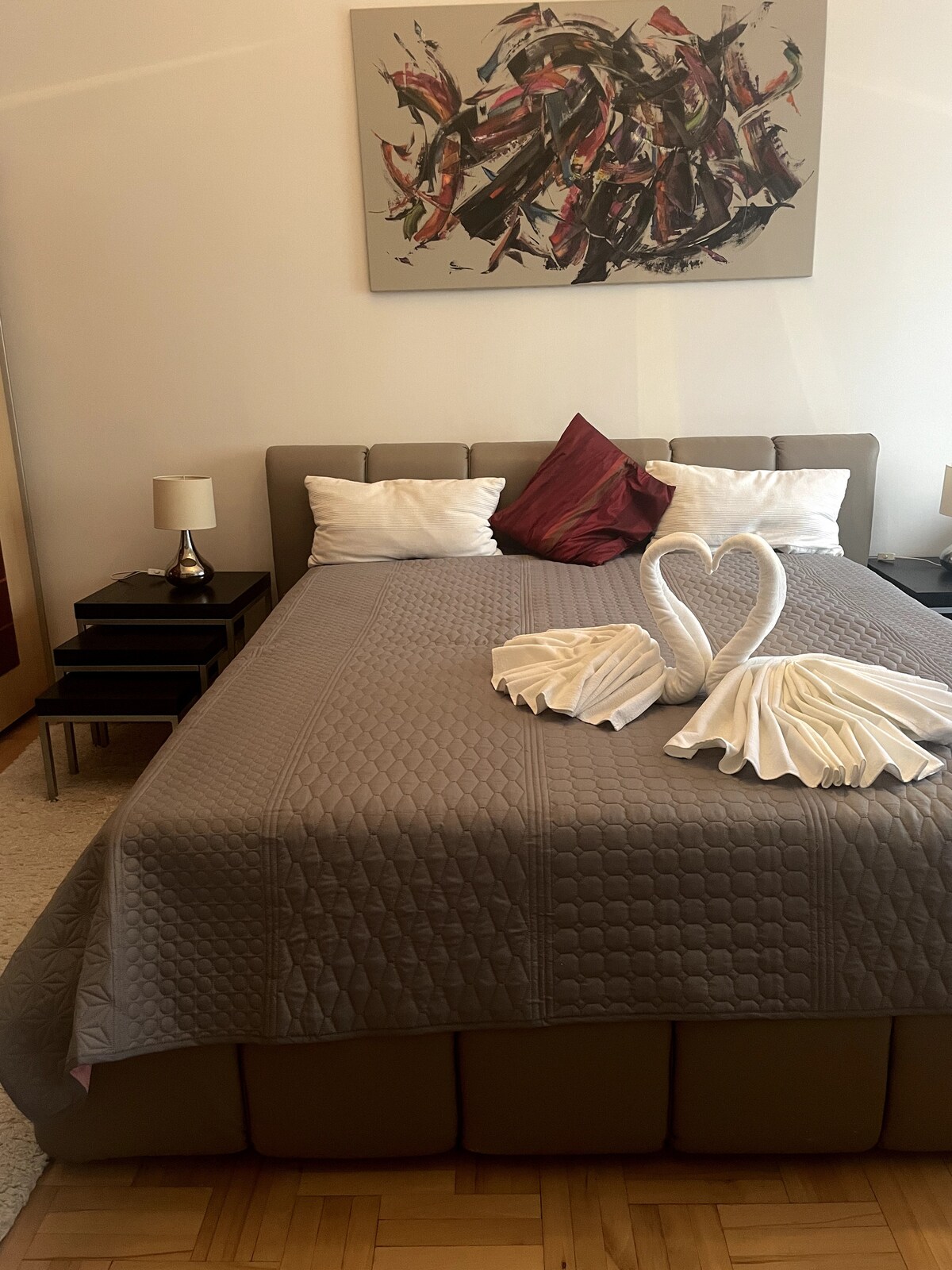 A spacious bed is adorned with a grey quilt and decorative pillows, including a vibrant red accent pillow. Beside the bed are two nightstands with lamps, providing soft lighting. An artistic wall hanging adds a touch of color to the room.