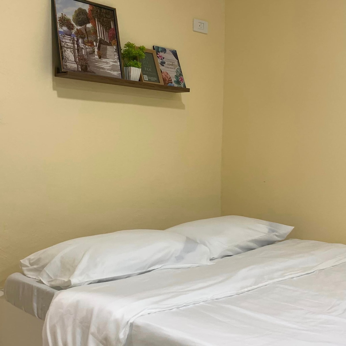 A cozy sleeping area with a spacious bed featuring fresh white linens. A wooden shelf is mounted on the wall above, displaying framed artwork and a small plant, providing subtle decor against the soft, warm walls.