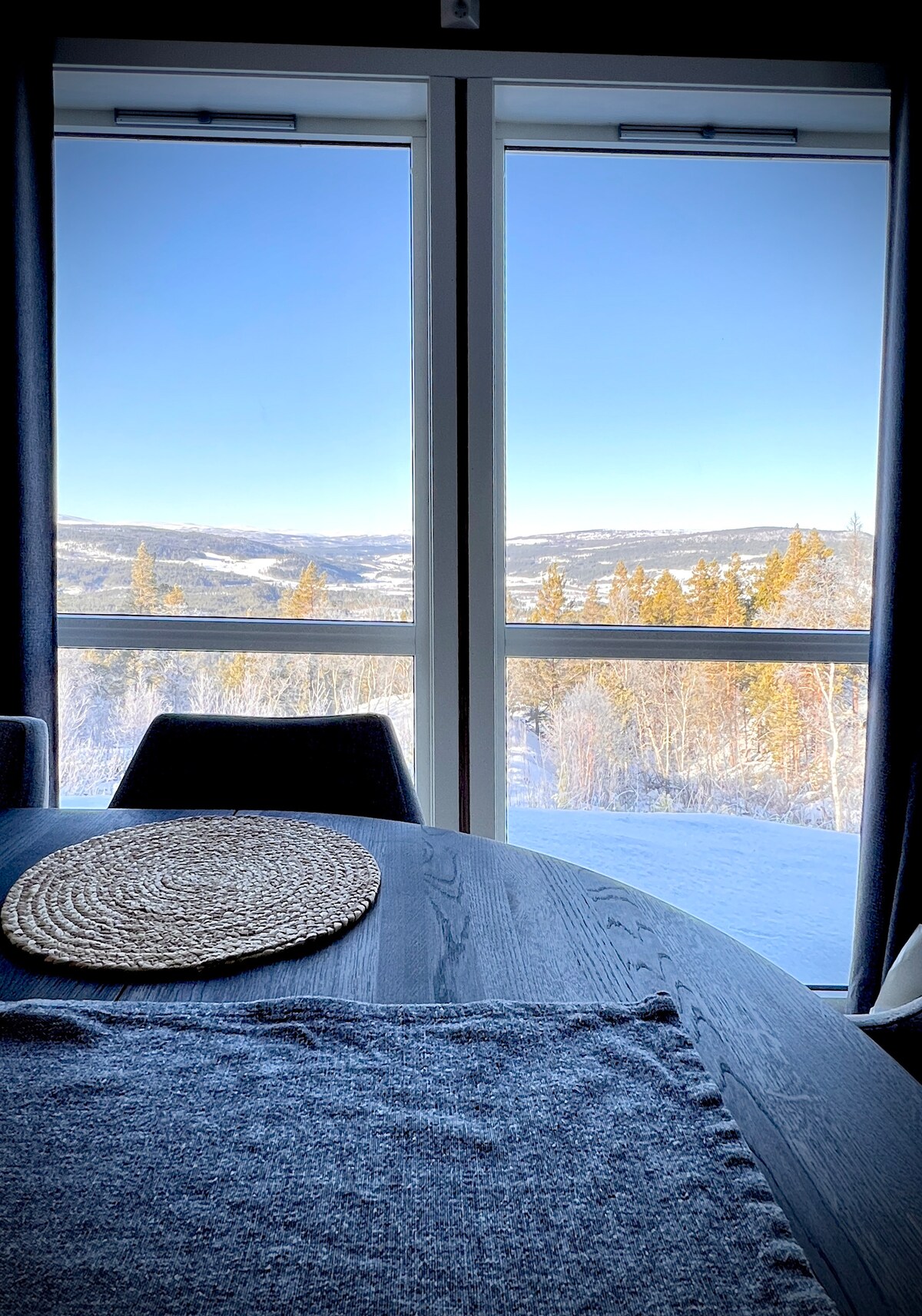 Large windows frame a winter landscape, revealing snowy hills and trees bathed in sunlight. A round woven placemat rests on a dark wooden table, highlighting the spacious dining area. Soft curtains drape alongside the windows, allowing natural light to fill the room.