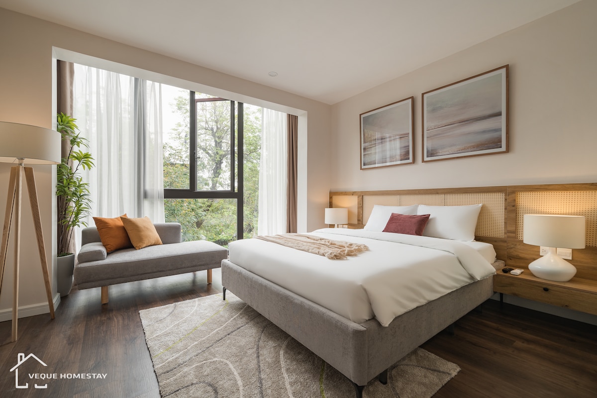 A serene sleeping area features a spacious bed against a light-toned wall, with soft natural light filtering through large glass windows adorned with sheer curtains. A cozy armchair is placed next to a small side table, and decorative artwork complements the calm decor.
