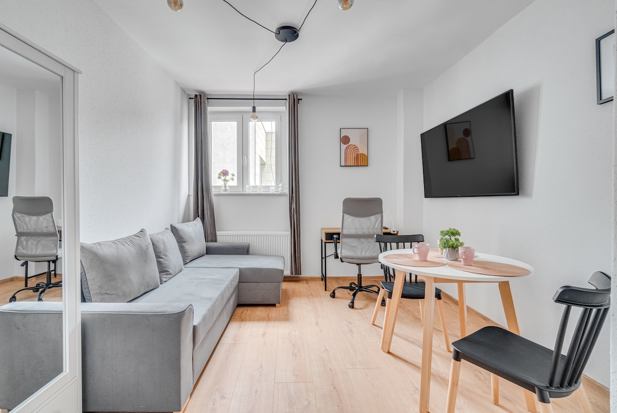 A modern living area features a sectional gray sofa and a round table set for two. A desk and chair are positioned opposite a wall-mounted television. Natural light enters through a window adorned with gray curtains, enhancing the room's functional layout.