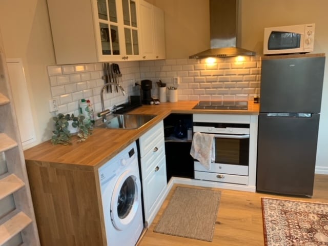 The kitchen area features modern cabinetry and appliances, including a refrigerator, stove, and microwave. A washing machine is integrated into the cabinetry, while a wooden countertop provides workspace. Clean white tiles complement the space, and a small area rug adds warmth.