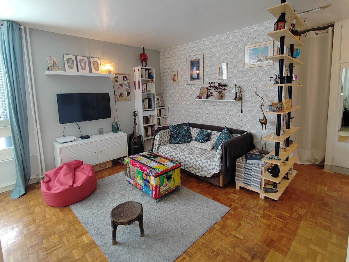 A spacious living area features a comfortable sofa adorned with colorful cushions. A vibrant, decorative coffee table stands on a soft area rug, while a television is mounted on the wall. Bookshelves filled with assorted items line one side of the room, and a bean bag chair adds a casual seating option.