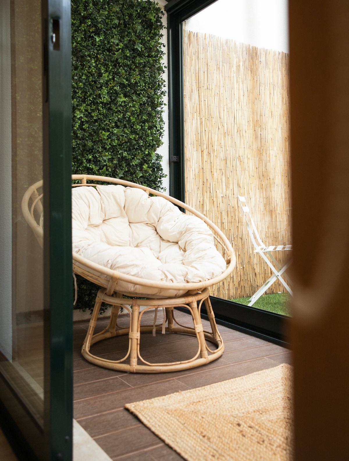 A cozy, circular rattan chair is positioned on a wooden floor, framed by large glass doors. A green, textured wall and a bamboo screen provide privacy, while a small table and chair are visible in the background, enhancing the outdoor ambiance.