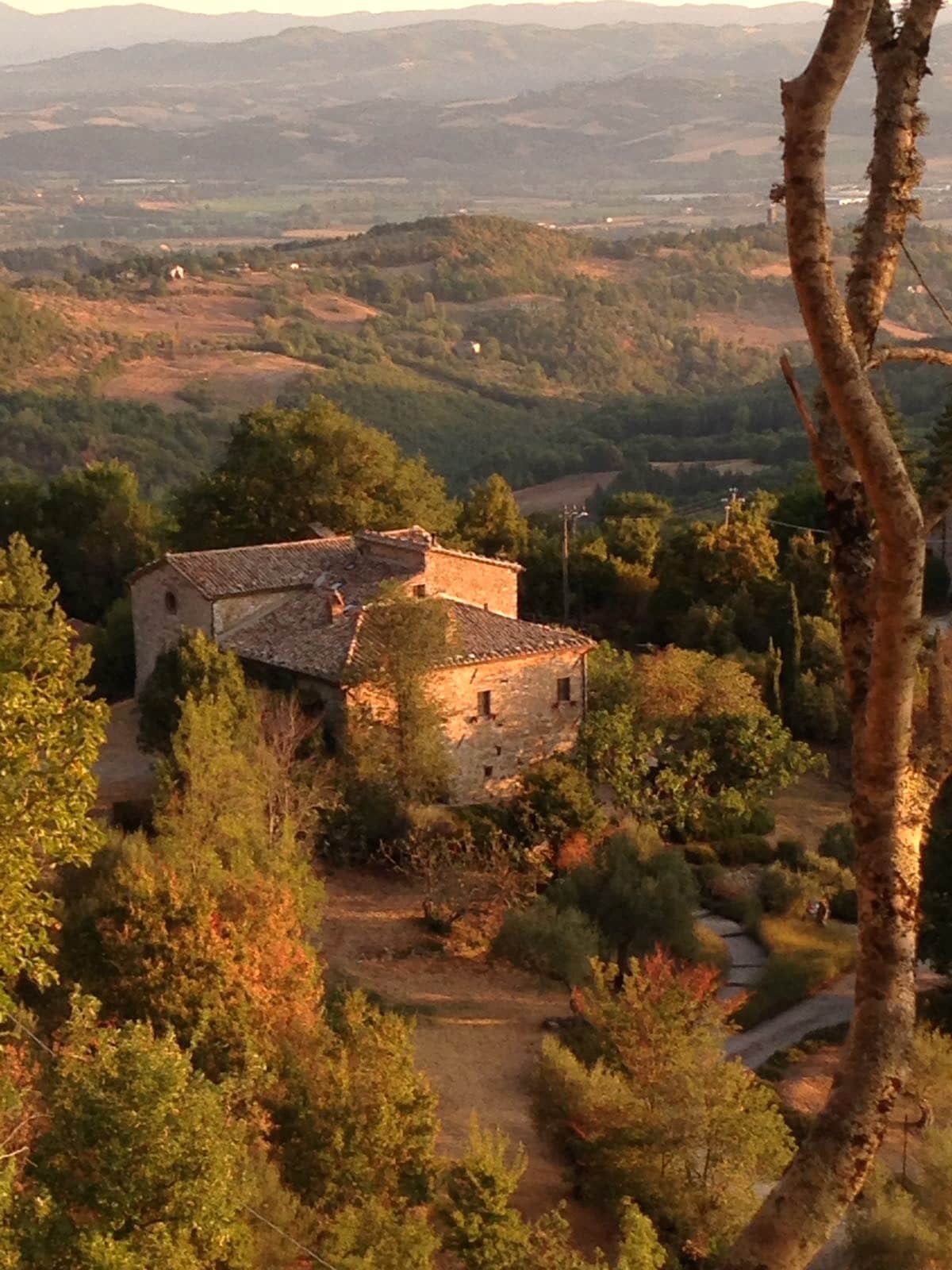 A charming stone house is nestled among rolling hills and trees, showcasing a rustic roof and traditional architectural features. The surrounding landscape includes fields and distant mountains, highlighted by warm earth tones and the golden light of sunset.
