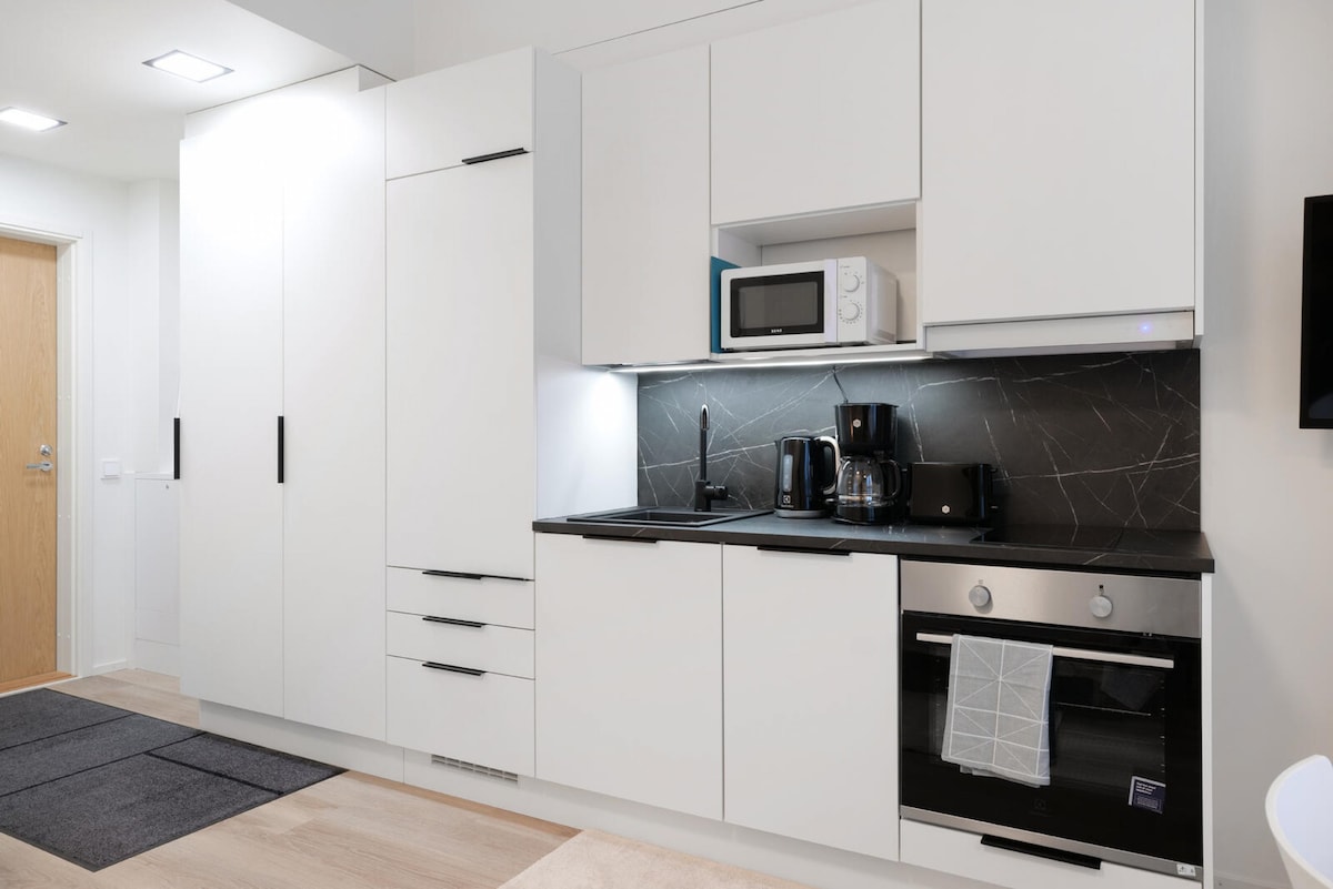 A modern kitchen features sleek white cabinetry and a dark stone countertop. Kitchen appliances include a microwave, coffee maker, and kettle, all presented with minimalistic elegance. The space is well-lit, enhancing the clean lines and functional design of the kitchen.