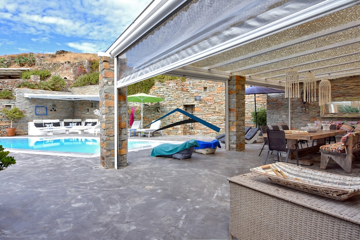 An outdoor space features a swimming pool with lounge chairs nearby and a shaded dining area. Stone walls provide a rustic backdrop, while various seating options enhance comfort. A covered area protects against the elements, allowing for relaxation and gatherings. An inviting atmosphere is created by the harmonious design.