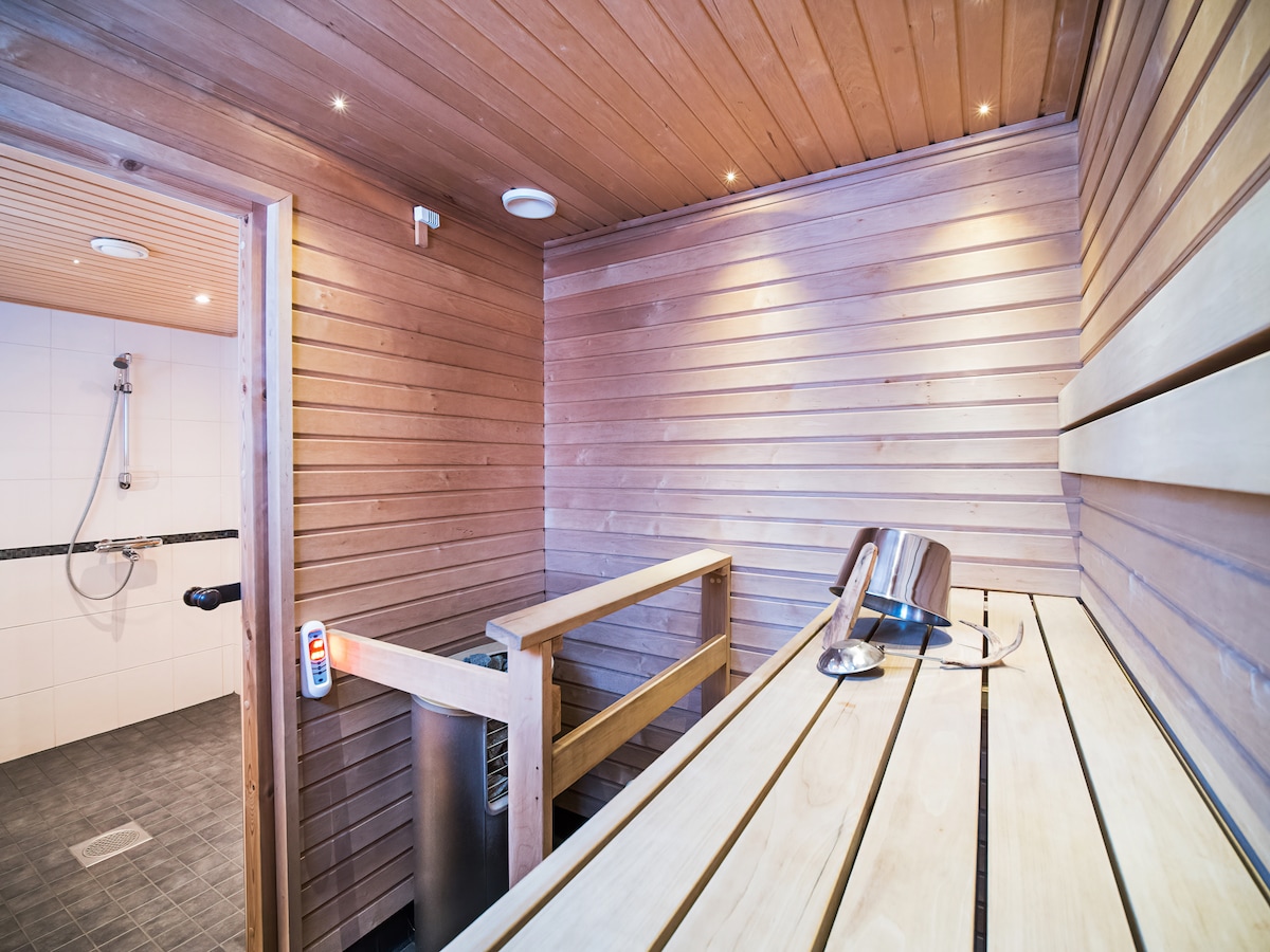 A serene sauna area is presented, featuring warm wooden walls and benches. A sleek shower is visible through an opening, while a small heater is placed beside the seating. Soft lighting enhances the calming atmosphere, creating a perfect space for relaxation.
