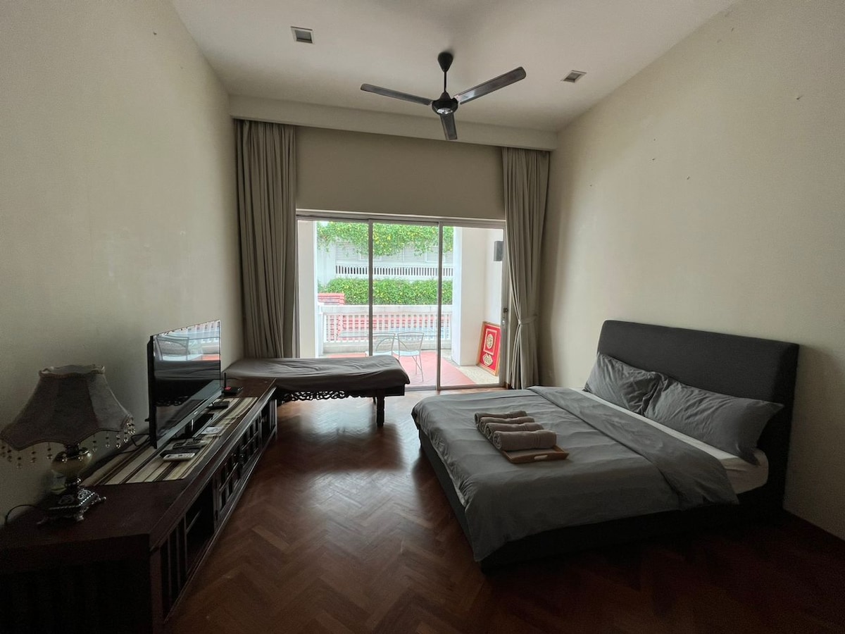 A spacious sleeping area is highlighted by a large bed covered with a soft grey blanket. A comfortable seating area is positioned beside a large glass window, allowing natural light to fill the room. A flat-screen television is mounted on a cabinet against the wall.