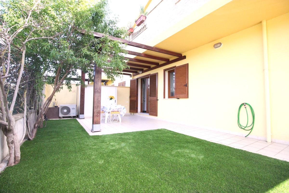 A well-maintained outdoor area features a manicured green lawn, with a covered patio that includes a table and chairs. The bright walls of the adjoining building provide a cheerful backdrop, and a wall-mounted air conditioning unit is visible. Access to the interior is through wooden shuttered doors.