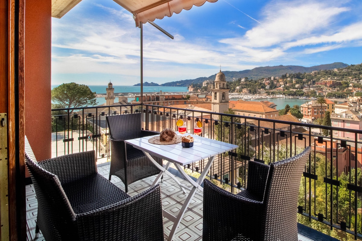 A balcony view showcases the scenic coastline and charming town. A table is set with refreshments, surrounded by four black wicker chairs. The tranquil setting is complemented by clear skies and gentle hills in the background, inviting relaxation.