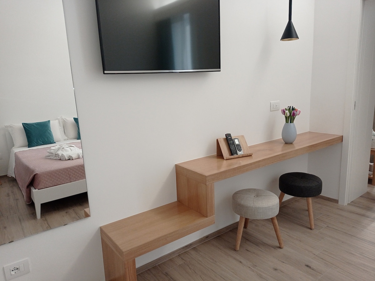 A minimalist space features a wooden console with two stools, one light and one dark, situated below a wall-mounted television. A small vase with blooming flowers adds a touch of color. Reflections of the bedroom area are visible in the nearby mirror.